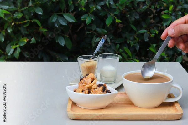 Fototapeta Part of hand holding spoon to stir coffee and there are cookies, sugar and milk together on wooden tray. Garden atmosphere.