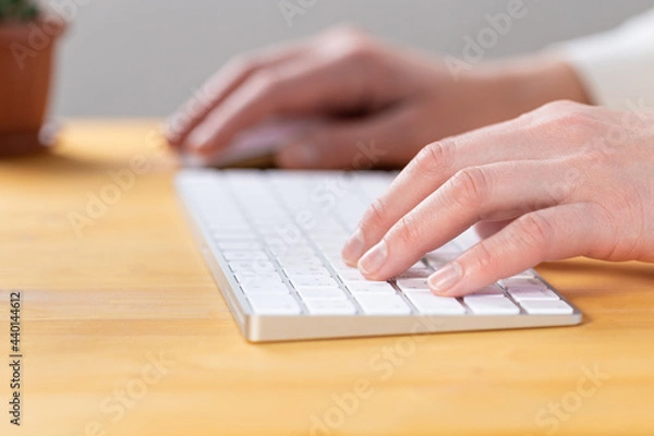 Fototapeta An image of a woman's hand is typing on a white keyboard on a wooden table. Selective focus