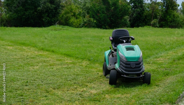 Fototapeta Green grass trimming with lawn mower. Tractor mowing grass in a public park. A dirty lawn tractor after mowing the lawn stands on the meadow. Professional lawn mower cuts the grass.