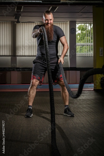 Fototapeta Strong young bearded man working out with battle ropes a gym. Determined guy using battle rope while doing physical training. Hard workout