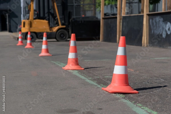 Fototapeta Bright orange traffic cones standing in a row on gray asphalt in the city. Road works. Selective focus. Hyster forklift in the background. Traffic cone with white and orange stripes near car outdoors