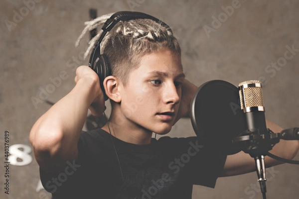 Obraz Stylish attractive guy with dreadlocks is recording a song in the studio. A young singer in black studio headphones stands in front of a microphone. Generation Z young talent