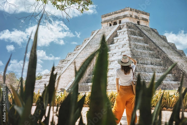 Fototapeta A young woman tourist in a hat stands against the background of the pyramid of Kukulcan in the ancient Mexican city of Chichen Itza. Travel concept.Mayan pyramids in Yucatan, Mexico