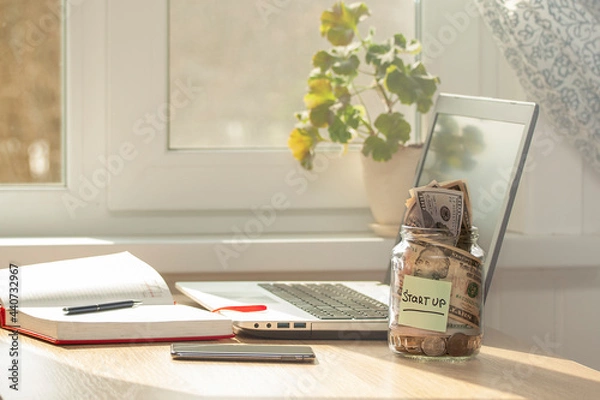 Fototapeta A glass jar with STARTUP written on it, with coins and banknotes inside stands on a desk.The concept is funding and promoting an innovative idea. Laptop, smartphone, diary on the desk.