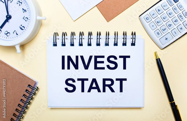 Fototapeta On a light background, a white alarm clock, a calculator, a pen and a notebook with the text INVEST START. Flat lay