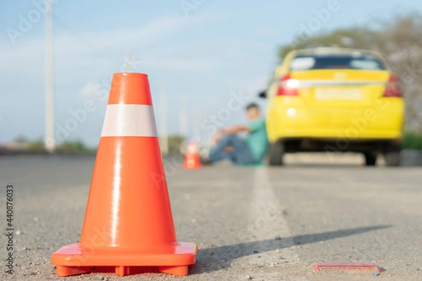 Fototapeta Traffic cone behind a broken down taxi and its driver in Latin America. Orange traffic sign and a damaged yellow car. Horizontal image.