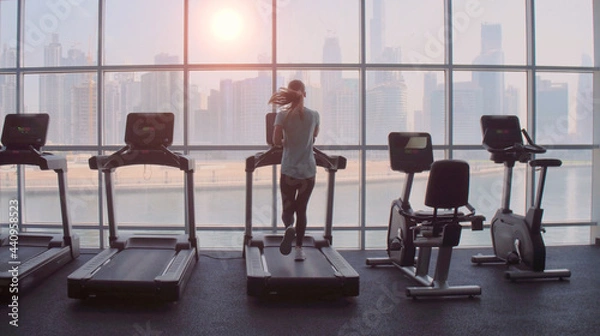 Fototapeta Woman running on treadmill at a panoramic window with city view at the sunrise.