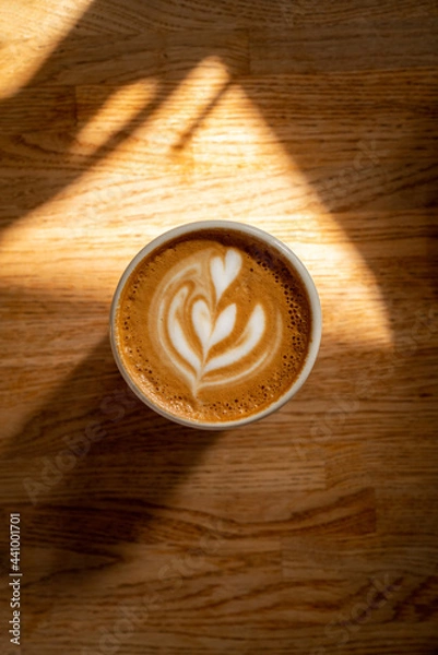 Fototapeta Coffee cup on wooden table. Fresh cappuccino with beautiful latte art on brown background with shadow and sun spots. Coffee break. Atmospheric photo. Good morning drink. Top view.