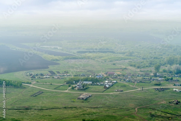 Fototapeta View from the mountain to the Russian village with a church and fields through a foggy haze. Photo taken near Andreevka village, Orenburg region, Russia