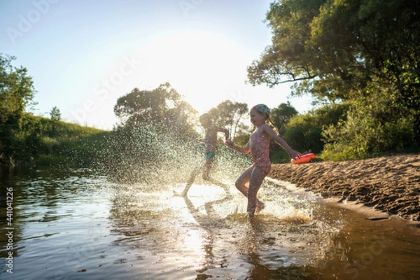 Fototapeta Happy children run from the shore into the water. Summer children's vacation on shore of a lake or river. boy and girl jump into water, swim and splash around at sunset. Active holidays. Dynamic image