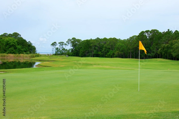 Fototapeta Lush golf course green with yellow flag in the hole and fairway with water trap, sand traps and tree line in central Florida on a crisp summer morning.