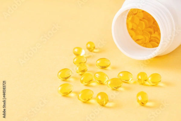 Fototapeta Transparent softgel capsules with fat-soluble vitamins and open plastic bottle on yellow background. Close-up, copy space.