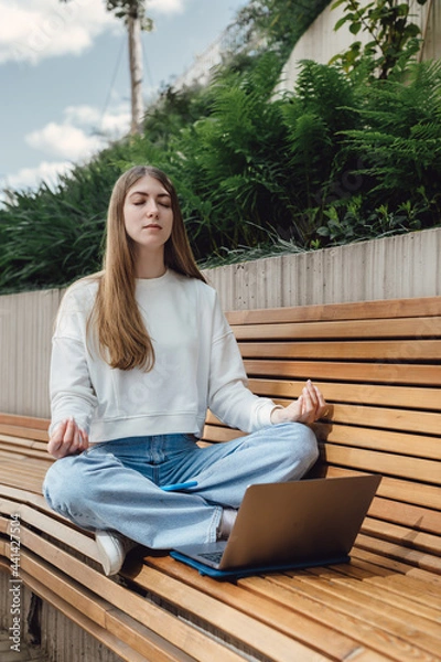 Fototapeta A young student girl sits in an urban park in the city and meditates during a break before studying science. University campus, education and study in the open air.
