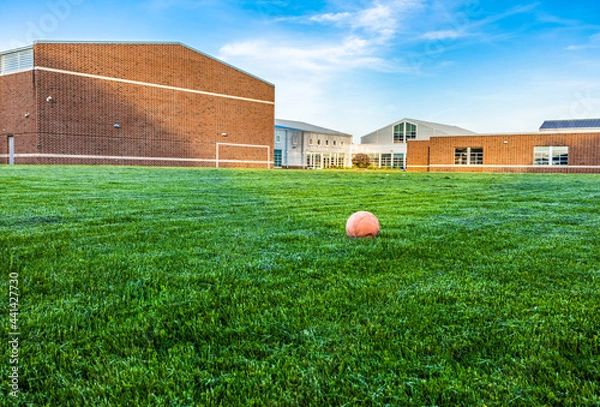 Fototapeta Old and deflate orange football on green turf with sunlight at school building background, depth of field. Outdoor recreation space for students. Landscape view of empty soccer field and unused ball.