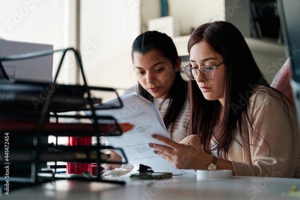 Obraz two young latina business women talking and working together in office, using documents and office supplies, teamwork concept