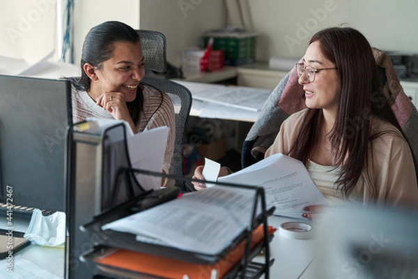 Obraz two young latina business women talking, laughing and working together in the office, using documents and office supplies, concept of teamwork.