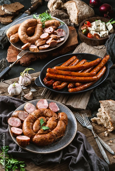 Fototapeta Cold meat assortment with rustic sausages on a plate, fried sausages in pan, garlic and fresh herbs on wooden background. Meat appetizer, top view
