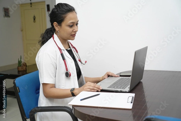 Obraz A south Indian female doctor in 30s with laptop and prescription board with white coat and red stethoscope in white background.Online medical consultation