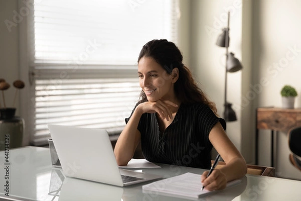Fototapeta Smiling Caucasian woman sit at desk in office work online on laptop consult client or customer. Happy successful businesswoman busy with computer job at workplace, consider brainstorm with documents.