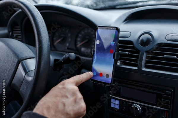 Fototapeta The driver in the car uses a smartphone mounted in a car holder on a torpedo close-up. blurred focus