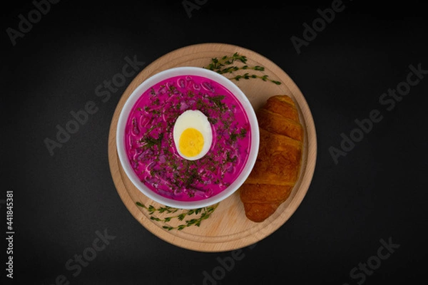 Fototapeta Summer cold beetroot soup (choldnik) with boiled egg served in a bowl on a wooden board with a croissant. Isolated black background. Top view.