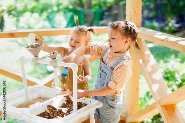 Fototapeta Happy sister kids play with sand and water in sensory baskets on the outdoor sensory table, sensory early development, montessori. Toddler and Big Sister Play with Sand, Soft Focus