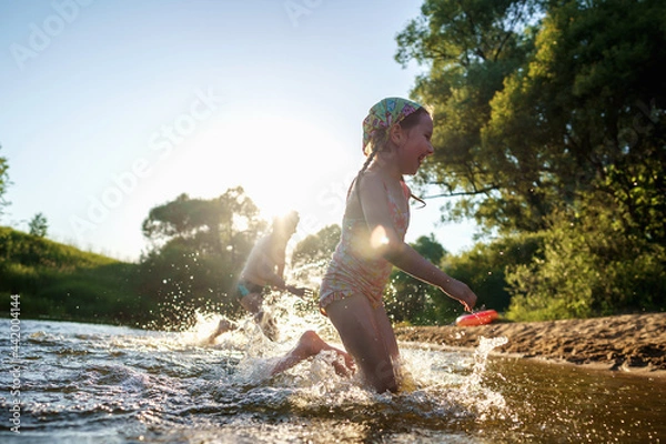 Fototapeta Happy children run from the shore into the water. Summer children's vacation on shore of a lake or river. boy and girl jump into water, swim and splash around at sunset. Active holidays. Dynamic image