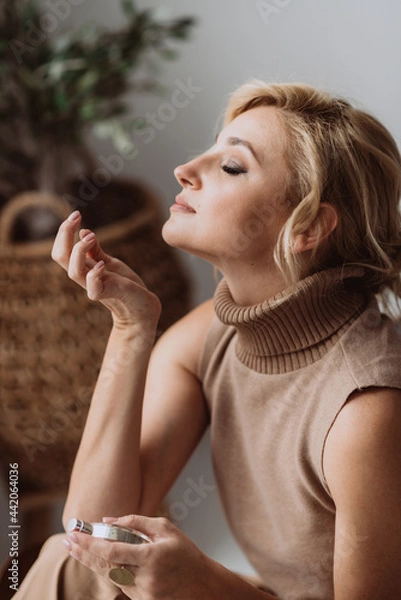 Fototapeta Young beautiful woman holds a bottle of perfume in her hands and inhales while enjoying the fragrance from her wrist. Soft selective focus.