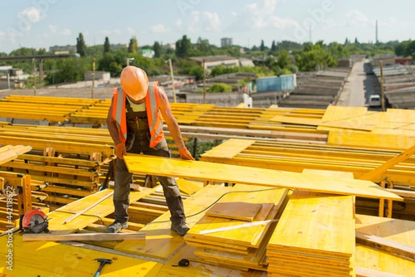 Fototapeta A worker in a safety helmet assembles a formwork from wooden panels