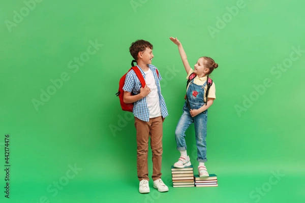 Fototapeta happy teenage schoolboy with a backpack, measuring the height of a little girl in loose clothes, standing on a stack of books on a green background. The concept of development and growth of education.