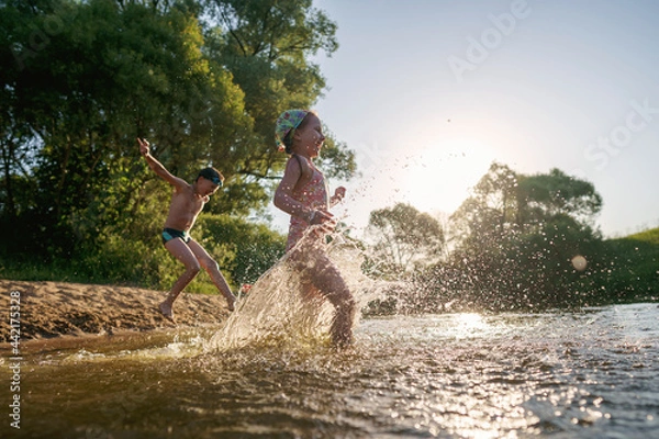 Fototapeta Happy children run from the shore into the water. Summer children's vacation on shore of a lake or river. boy and girl jump into water, swim and splash around at sunset. Active holidays. Dynamic image