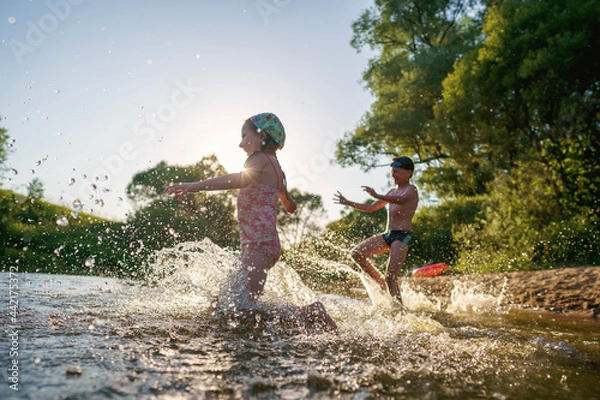 Fototapeta Happy children run from the shore into the water. Summer children's vacation on shore of a lake or river. boy and girl jump into water, swim and splash around at sunset. Active holidays. Dynamic image