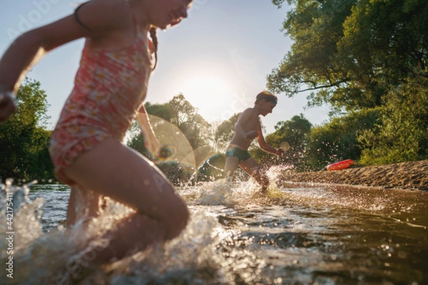 Fototapeta Happy children run from the shore into the water. Summer children's vacation on shore of a lake or river. boy and girl jump into water, swim and splash around at sunset. Active holidays. Dynamic image