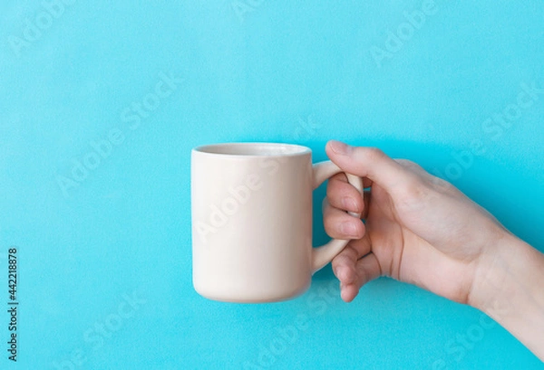 Obraz Human hand holding big ceramic beige mug above light blue background with copy space. Mug mock up for your design
