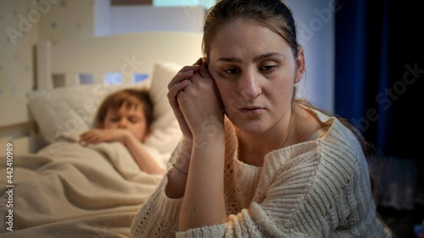 Fototapeta Upset and stressed mother crying next to bed of her little son sleeping at night. Concept of lonely mother, family problems and maternal depression