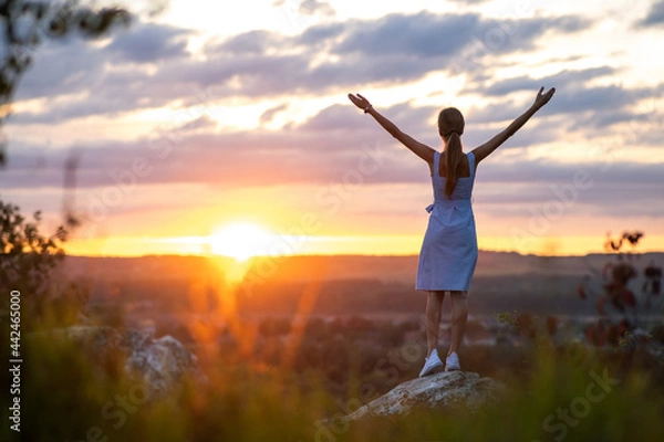Fototapeta Dark silhouette of a young woman standing with raised up hands on a stone enjoying sunset view outdoors in summer.
