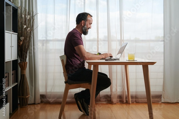 Fototapeta smiling middle-aged man working at home with his laptop computer on a wooden table next to a large window