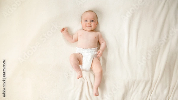 Fototapeta Top view of little baby boy in diapers lying on soft white blanket on bed.