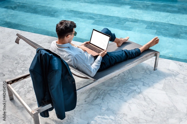 Fototapeta Young businessman resting on sunbed enjoying summertime in resort spa near swimming pool while working remotely at laptop computer connected to wireless internet.
