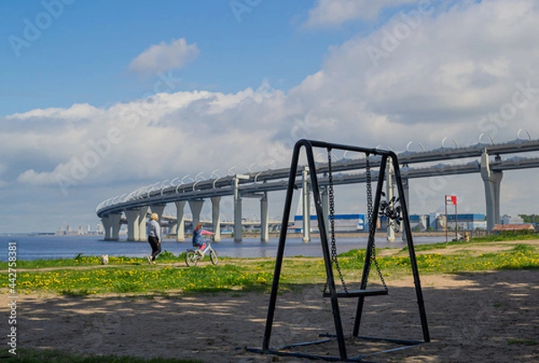 Fototapeta 
provincial village is swayed by the wind against the background of large round highway bridge in the blue sea and sky with clouds. People, leisure, concept past and future building world