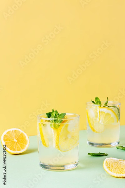 Fototapeta Two glasses of ice lemon water with mint leaves on a yellow and green background. Fresh summer drink with soda.