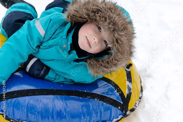 Obraz A boy in a turquoise overalls with a hood lies on a tubing in the snow and smiles. People, lifestyle concept