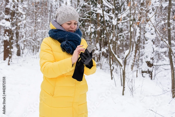 Obraz A woman in a bright yellow jacket holds a phone in her hands, smiles and walks in a snowy forest. People, lifestyle concept