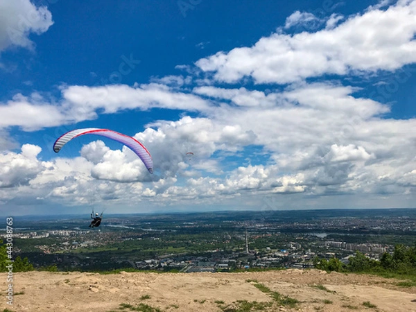 Fototapeta Paraglider taking off from the edge of the mountain over a city. Blue sky with clouds. Flying with a colordul parachute. Concept of freedom, relax, paragliding in the air