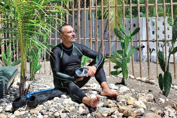 Fototapeta A man in a diving suit sits on the seashore and looks into the distance