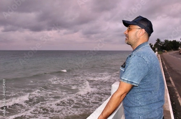 Fototapeta A man standing on a bridge against the backdrop of a stormy sea and gray sky and looking into the distance