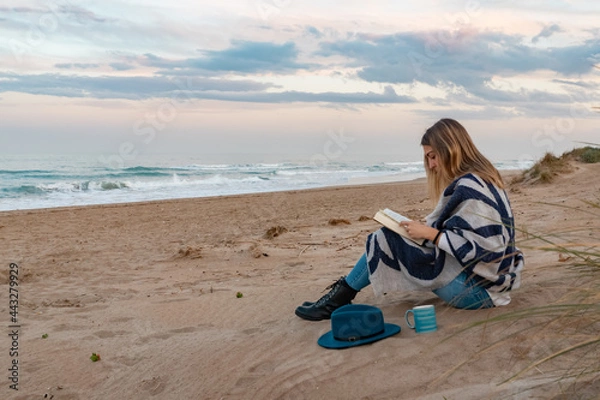 Fototapeta a girl with a blue hat is taking a coffee watching the sea while is reading a book