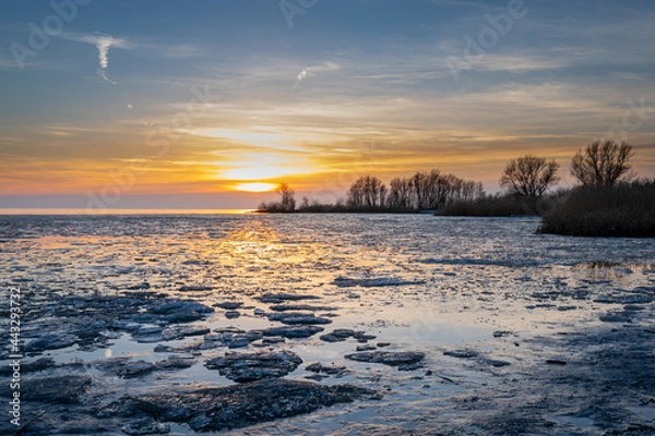 Fototapeta Sunset on ice lake, Melting ice on the water of a lake during sunset light cloudy blue orange sky and trees on background