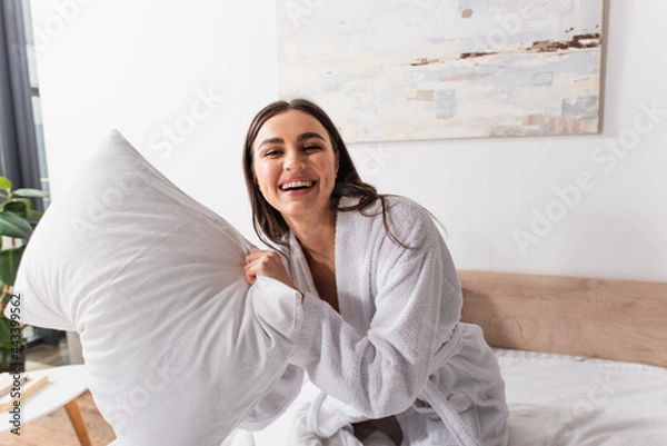 Obraz playful young woman in bathrobe holding pillow and laughing while looking at camera