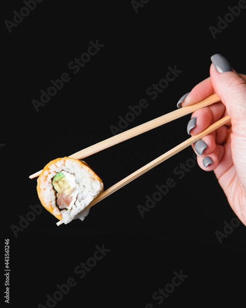 Fototapeta Hand holding sushi roll using chopsticks on black background. Cheddar cheese sushi roll over black